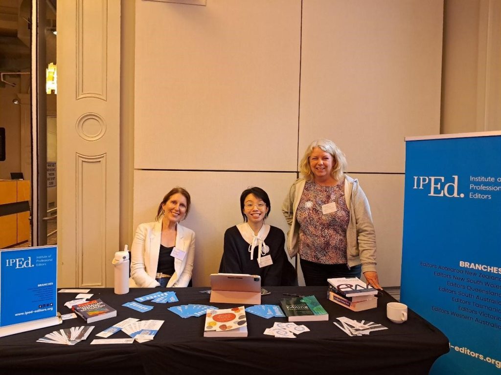 Three woman sit behind a table. Beside them is an IPEd pop up banner. On the table are books, bookmarks and stickers promoting IPEd and editing.