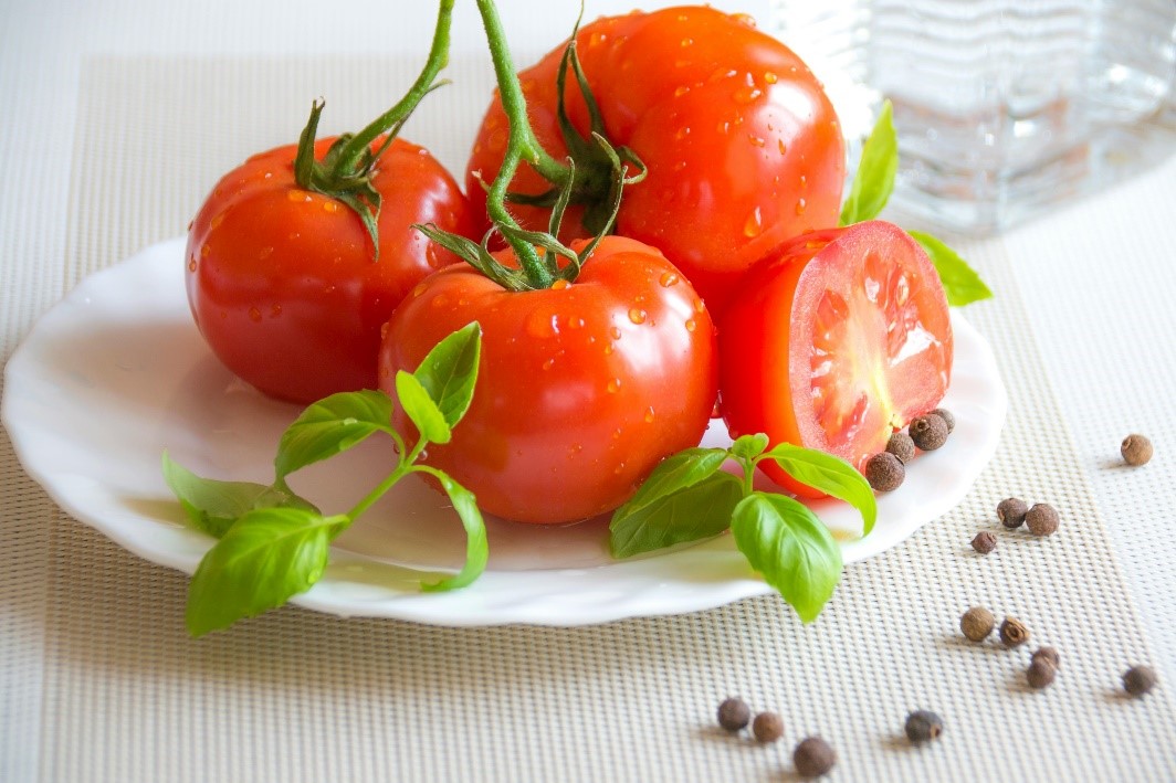 Three and a half tomatoes with sprigs of basil on a white plate. The whole tomatoes have their stalks attached. A few peppercorns are sprinkled on the plate and more are on the table to the right of it.