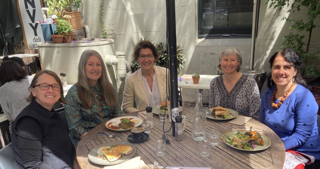 A group of people sit around a timber table at a cafe. They are outside. 