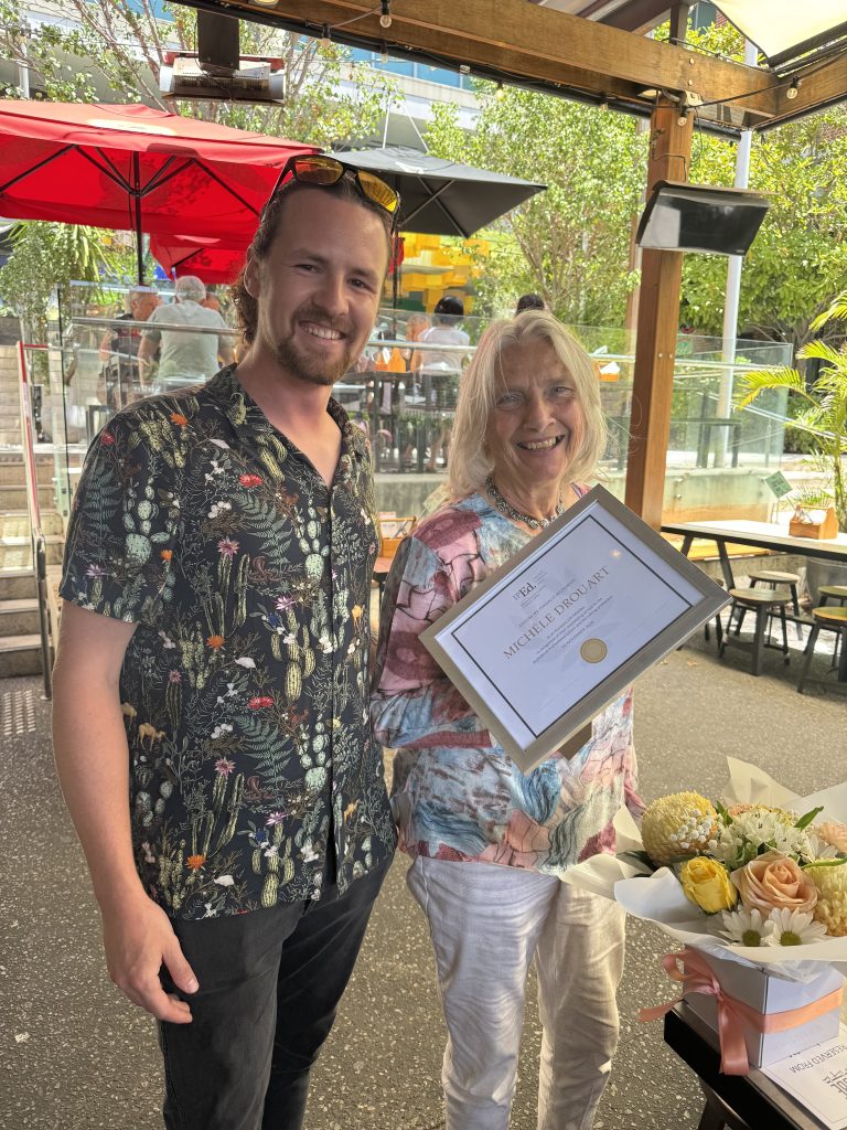 A man and a woman stand next to each other at an outdoor cafe. They are both smiling. She holds a certificate and there is a bunch of flowers on a table in front of them.